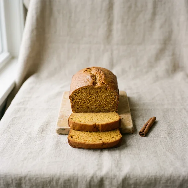 A loaf of pumpkin bread on a small wooden board with two slices in front and a cinnamon stick beside it